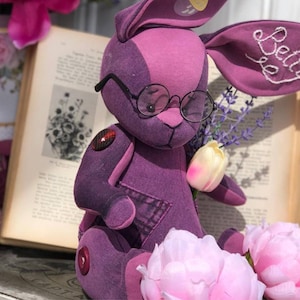 Könnte beinhalten: Ein lila Plüschhase mit runder Brille, der einen Lavendelzweig und eine Tulpe hält. Der Hase hat den Namen "Betty" auf einem Ohr gestickt. Ein Vintage-Buch und rosa Pfingstrosen sind im Hintergrund.