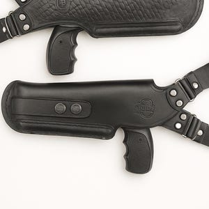 May include: Two black leather shoulder holsters. One has a textured, embossed pattern, and the other a smooth finish. Both have molded grips and adjustable straps with metal hardware. The holsters are designed to be worn across the chest.