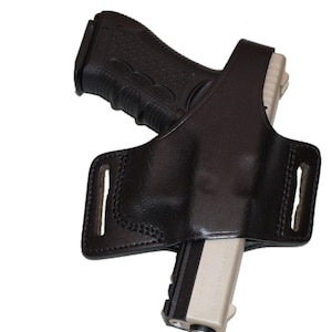 Puede incluir: Funda de cuero negro diseñada para sujetar una pistola. La funda tiene una forma moldeada para sujetar el arma de fuego de forma segura. El arma está parcialmente insertada, con la empuñadura y la corredera visibles. La funda tiene trabillas.