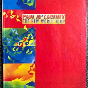 Op de afbeelding: Een rechthoekig tourprogramma met een felrode omslag met de tekst PAUL MCCARTNEY THE NEW WORLD TOUR in een geel kader. De linkerkant toont drie gestapelde panelen met abstracte geometrische vormen in blauw, groen, geel en rood.