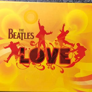 May include: A square, yellow album cover with the text "THE BEATLES" in brown letters. The word "LOVE" is in large red letters. Silhouettes of the Beatles are in red, with a yellow and orange background.