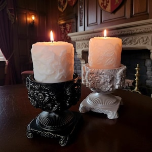 Peut inclure: Deux bougies piliers allumées dans des supports ornés. Une bougie est blanche, l'autre est pêche. Le support noir a un motif floral, tandis que l'autre est argenté. Les bougies sont sur une surface en bois sombre.