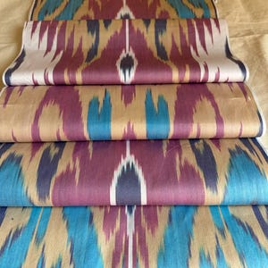 May include: Several rolls of patterned fabric in teal, purple, gold, and white. The fabric displays a repeating ikat design with vertical stripes and geometric shapes. The rolls are neatly stacked, highlighting the intricate patterns and colours.