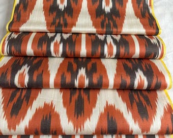 Può includere: Tessuto piegato con un motivo ikat ripetuto in tonalità arancione, marrone, bianco e nero. I bordi sono rifiniti con un bordo giallo. Il tessuto è probabilmente seta o un materiale simile.