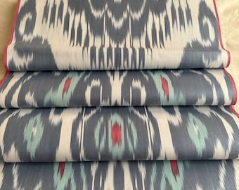 May include: Folded fabric with a repeating ikat pattern in shades of gray, white, red, and teal. The edges are finished with a thin pink border. The fabric is stacked, showcasing the detailed design.