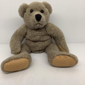 May include: A plush teddy bear in a seated position. The bear is a muted brown color with a black nose and eyes. The bear's paws are a lighter tan color. The bear is made of a soft, textured material.