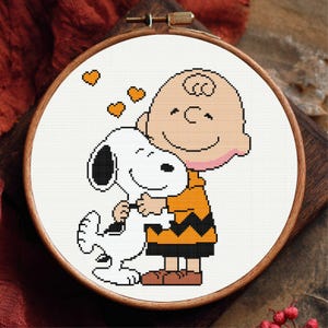 Puede incluir: Un bastidor de bordado de punto de cruz con una imagen de dibujos animados de Charlie Brown abrazando a Snoopy. Los personajes están en un cálido abrazo, Snoopy es blanco y negro, y Charlie Brown lleva una camisa naranja. Tres corazones naranjas flotan sobre ellos.