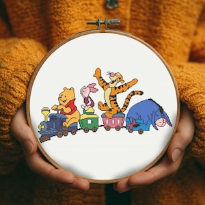 Puede incluir: Un bastidor de bordado de punto de cruz con Winnie the Pooh y sus amigos en un tren. Los personajes son Winnie the Pooh, Piglet, Tigger y Eeyore. Los vagones del tren son coloridos, sobre un fondo blanco.