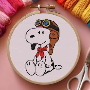 May include: Cross-stitch embroidery of Snoopy the dog wearing a brown aviator helmet and goggles, and a red bow tie. The design is set in a wooden embroidery hoop against a light pink background. Colourful embroidery floss and scissors are visible.