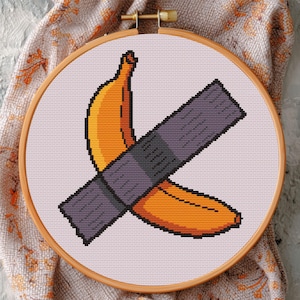 Könnte beinhalten: Kreuzstich-Stickerei einer Banane, die mit grauem Klebeband befestigt ist. Die Banane ist orange und gelb. Die Stickerei befindet sich in einem hölzernen Stickrahmen.