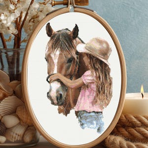 May include: Cross-stitch embroidery of a horse and a person in a wooden hoop. The horse is brown and white, and the person is wearing a pink shirt, blue jeans, and a hat. The embroidery is set against a white background.