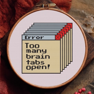Può includere: Ricamo a punto croce in un telaio di legno. Il disegno presenta un messaggio di errore del computer pixelato che recita "Error: Too many brain tabs open!" in testo nero su sfondo beige, con accenti rossi e blu.