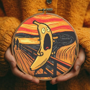 Pode incluir: Um bastidor de bordado em ponto cruz com uma paródia humorística de "O Grito" de Edvard Munch. A obra de arte substitui a figura por uma banana gritando contra um fundo vibrante laranja e preto. O bastidor de madeira é segurado nas mãos de uma pessoa.
