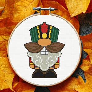 May include: A cross-stitch embroidery of a nutcracker soldier in a wooden hoop. The design features a green and gold hat with a red plume, a brown mustache, and a white beard. The background is white, and the hoop is set against a backdrop of autumn leaves.