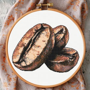 May include: Cross-stitch embroidery of three coffee beans in a wooden hoop. The beans are shades of brown and tan, with detailed shading. The embroidery is on white fabric, set against a patterned cloth.