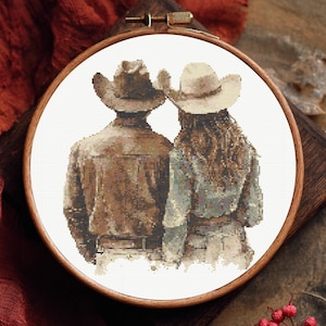 May include: Cross-stitch embroidery of a couple in cowboy hats and jackets, seen from behind. The artwork is framed in a round wooden hoop. The colour scheme features browns, beiges, and blues, giving a rustic feel to the piece.