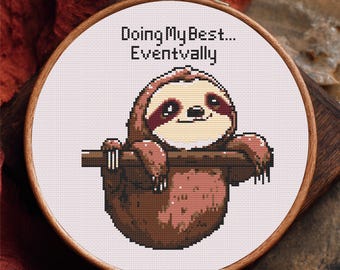 May include: Cross-stitch embroidery in a wooden hoop featuring a smiling sloth hanging from a branch. The text "Doing My Best... Eventually" is above the sloth. The sloth is brown and tan, with a white face and dark eyes.