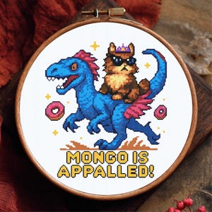 May include: Cross-stitch embroidery in a wooden hoop features a cat wearing sunglasses and a crown riding a blue dinosaur with pink wings. The text "MONGO IS APPALLED!" is stitched below the image, along with two donuts and gold stars.