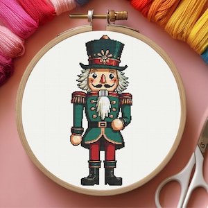 May include: Cross-stitch embroidery of a nutcracker soldier in a wooden hoop. The nutcracker is dressed in a green coat, red pants, and black boots. The background is white, and colorful embroidery floss surrounds the hoop.