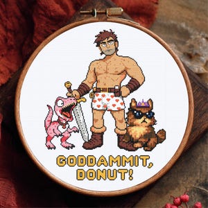 May include: A circular wooden embroidery hoop containing a cross-stitch design of a shirtless warrior in heart-patterned boxers holding a sword, a pink dinosaur, and a fluffy cat wearing sunglasses and a crown, with the text GODDAMMIT, DONUT!