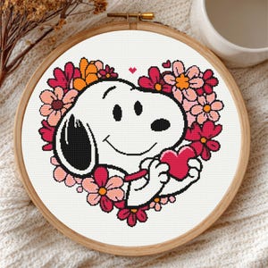 May include: A cross-stitch embroidery hoop featuring Snoopy holding a red heart, surrounded by a floral heart-shaped wreath. The design includes pink, red, and orange flowers. The hoop is set against a white fabric background.