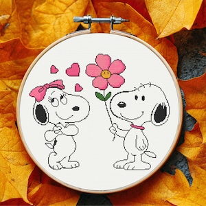Könnte beinhalten: Kreuzstich-Stickrahmen mit Snoopy und einer weiblichen Snoopy-Figur. Snoopy hält eine rosa Blume. Die weibliche Snoopy hat eine rosa Schleife und Herzen. Der Hintergrund besteht aus Herbstblättern.