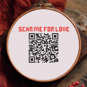 May include: A cross-stitch embroidery in a wooden hoop. The design features a black and white QR code with the red text "SCAN ME FOR LOVE" above it. The embroidery is on white fabric.