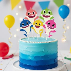 Op de afbeelding: Een blauwe ombre taart versierd met kleurrijke haaienthema toppers. De taart is versierd met vijf haai-uitsparingen in verschillende kleuren, waaronder roze, geel, blauw en groen. Ballonnen en feestvlaggen zijn op de achtergrond.