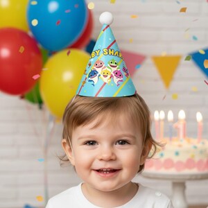 Op de afbeelding: Een lachend kind draagt een blauwe Baby Shark-verjaardagshoed met een witte pompon. De hoed heeft cartoonhaaien en de woorden "Baby Shark". Kleurrijke ballonnen en een verjaardagstaart met brandende kaarsen staan op de achtergrond.
