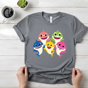 Op de afbeelding: Een grijs t-shirt met een kleurrijke afbeelding van vijf cartoon haaien in roze, groen, blauw en geel. De haaien hebben witte tanden en lachen. Het shirt wordt getoond op een wit houten oppervlak.