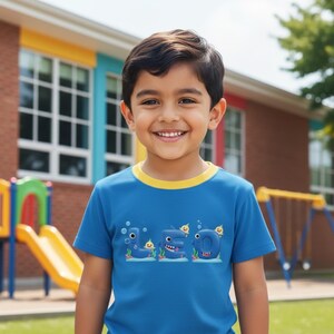 Op de afbeelding: Een kind draagt een blauw t-shirt met een gele kraag. Het shirt toont het woord "Sea" in een cartoonstijl, waarbij elke letter is ontworpen als een zeedier. De achtergrond omvat een schoolgebouw en een speeltuin.