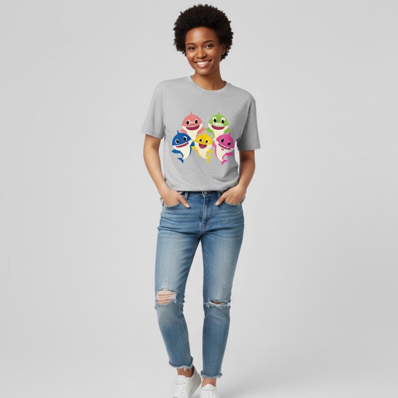 Op de afbeelding: Een lichtgrijs t-shirt met een kleurrijke afbeelding van zes cartoonhaaien. De haaien zijn in verschillende kleuren, waaronder roze, blauw, geel en groen. De persoon draagt een blauwe jeans met gescheurde knie&euml;n.