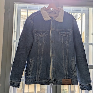 Può includere: Una giacca di jeans blu con colletto e fodera in sherpa color crema. La giacca ha quattro tasche frontali e una chiusura con bottoni. Un'etichetta in pelle marrone con la scritta "bunch" è cucita in basso a destra.