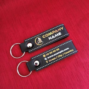 K&ouml;nnte beinhalten: Zwei schwarze Leder-Schl&uuml;sselanh&auml;nger mit silbernen Schl&uuml;sselringen. Der obere Schl&uuml;sselanh&auml;nger tr&auml;gt die wei&szlig;en Worte "COMPANY NAME" und ein goldenes Logo. Der untere Schl&uuml;sselanh&auml;nger enth&auml;lt Kontaktinformationen, einschlie&szlig;lich Telefonnummer, Website und Adresse.