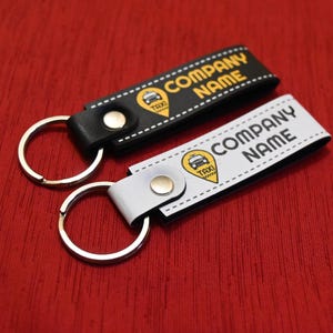 K&ouml;nnte beinhalten: Zwei Schl&uuml;sselanh&auml;nger mit Taxi-Motiv. Einer ist schwarz, der andere wei&szlig;. Beide haben einen silbernen Schl&uuml;sselring und die Worte "COMPANY NAME" in fetter Schrift. Ein gelbes Taxi-Symbol ist ebenfalls vorhanden.