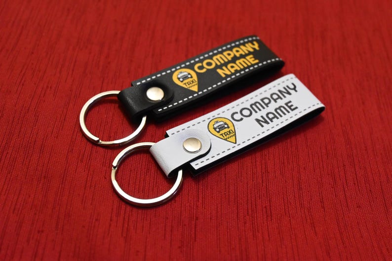 K&ouml;nnte beinhalten: Zwei Schl&uuml;sselanh&auml;nger mit Taxi-Motiv. Einer ist schwarz, der andere wei&szlig;. Beide haben einen silbernen Schl&uuml;sselring und die Worte "COMPANY NAME" in fetter Schrift. Ein gelbes Taxi-Symbol ist ebenfalls vorhanden.