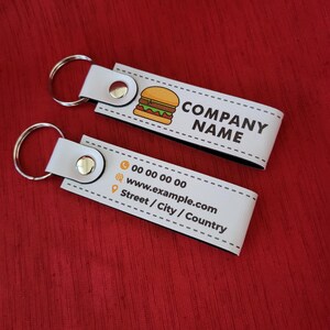 K&ouml;nnte beinhalten: Zwei wei&szlig;e Leder-Schl&uuml;sselanh&auml;nger mit silbernen Schl&uuml;sselringen. Jeder Schl&uuml;sselanh&auml;nger hat einen Druckknopfverschluss und ein gedrucktes Design. Einer zeigt eine Burger-Grafik und die Worte "COMPANY NAME". Der andere hat Kontaktinformationen.