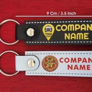 K&ouml;nnte beinhalten: Zwei Schl&uuml;sselanh&auml;nger, einer schwarz und einer wei&szlig;, mit silbernen Ringen. Der schwarze Schl&uuml;sselanh&auml;nger hat eine Taxi-Grafik und die Worte "COMPANY NAME". Der wei&szlig;e Schl&uuml;sselanh&auml;nger zeigt eine Pizza-Grafik und die Worte "COMPANY NAME". Die Schl&uuml;sselanh&auml;nger sind 9 cm lang und 2,5 cm breit.