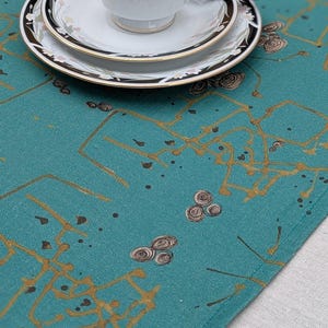 Könnte beinhalten: Ein weißes Tassen- und Untertassen-Set mit schwarzem und goldenem Rand, platziert auf einem passenden Teller. Das Set steht auf einem türkisfarbenen Tischläufer mit goldenen abstrakten Designs und braunen floralen Akzenten. Ein schwarzer Löffel liegt auf der Untertasse.