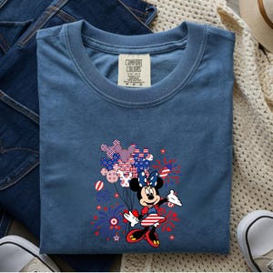 K&ouml;nnte beinhalten: Ein blaues T-Shirt mit einer Grafik von Minnie Maus in einem patriotischen rot-wei&szlig;-blauen Outfit. Sie h&auml;lt ein B&uuml;ndel Luftballons mit Sternen- und Streifenmuster, umgeben von Illustrationen roter und blauer Feuerwerke.