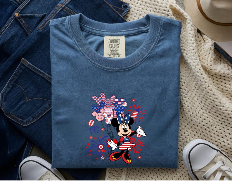 K&ouml;nnte beinhalten: Ein blaues T-Shirt mit einer Grafik von Minnie Maus in einem patriotischen rot-wei&szlig;-blauen Outfit. Sie h&auml;lt ein B&uuml;ndel Luftballons mit Sternen- und Streifenmuster, umgeben von Illustrationen roter und blauer Feuerwerke.