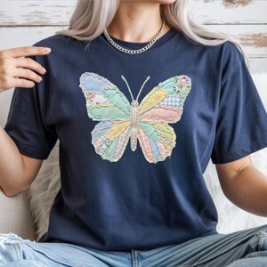 May include: Navy blue t-shirt featuring a colorful butterfly design. The butterfly is made of patchwork fabric in pastel colors, including blue, pink, yellow, and green. The butterfly has a detailed, textured appearance, and the shirt is a casual style.