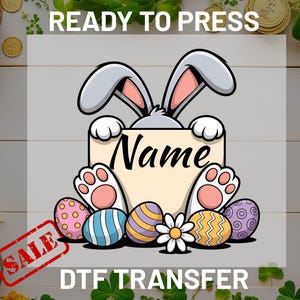 May include: A DTF transfer design featuring a cartoon bunny holding a sign with the word "Name". The design includes Easter eggs and a flower. The words "READY TO PRESS" and "SALE" are also visible.
