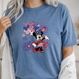 K&ouml;nnte beinhalten: Ein blaues T-Shirt mit kurzen &Auml;rmeln und einer Grafik einer Zeichentrickmaus, die ein patriotisches Kleid und eine Schleife tr&auml;gt. Das Design umfasst rot-wei&szlig;-blaue Luftballons in Form von Mausohren sowie Feuerwerksdarstellungen.