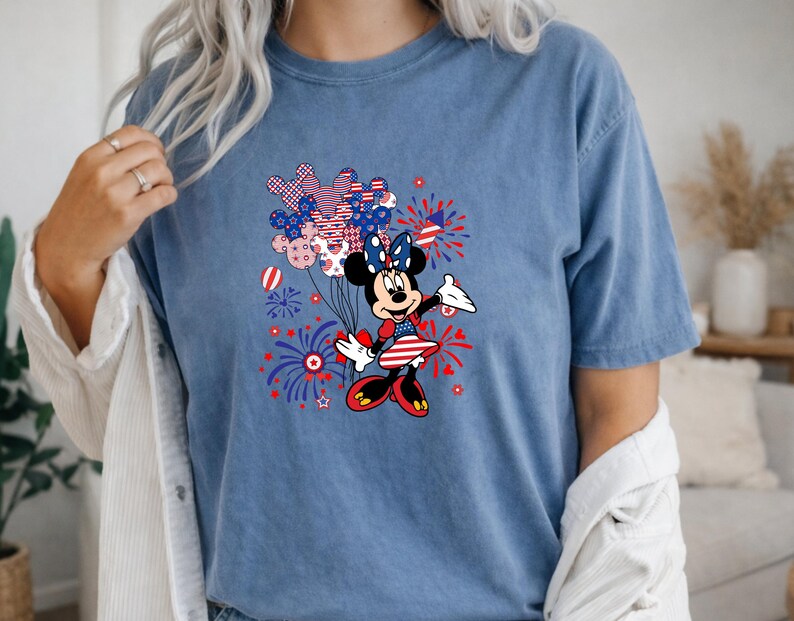 K&ouml;nnte beinhalten: Ein blaues T-Shirt mit kurzen &Auml;rmeln und einer Grafik einer Zeichentrickmaus, die ein patriotisches Kleid und eine Schleife tr&auml;gt. Das Design umfasst rot-wei&szlig;-blaue Luftballons in Form von Mausohren sowie Feuerwerksdarstellungen.