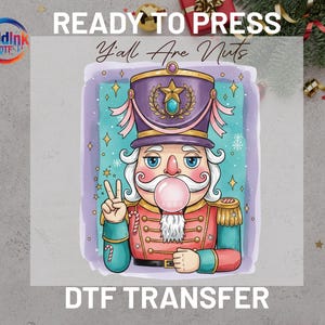 May include: A colorful DTF transfer featuring a cartoon nutcracker soldier with a bubblegum bubble. The nutcracker wears a purple hat with a gold star and a red and gold uniform. Text reads "READY TO PRESS" and "Y'all Are Nuts".
