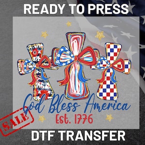 May include: A DTF transfer design featuring three crosses with patriotic bow accents. The crosses are adorned with red, white, and blue patterns, with the text "God Bless America Est. 1776". The word "SALE" is stamped on the bottom left.