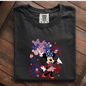 K&ouml;nnte beinhalten: Ein anthrazitfarbenes T-Shirt mit einer Grafik von Minnie Maus in einem rot-wei&szlig;-blauen Outfit. Sie h&auml;lt ein B&uuml;ndel Mickey-f&ouml;rmiger Luftballons mit patriotischen Mustern, umgeben von Illustrationen roter und blauer Feuerwerke.