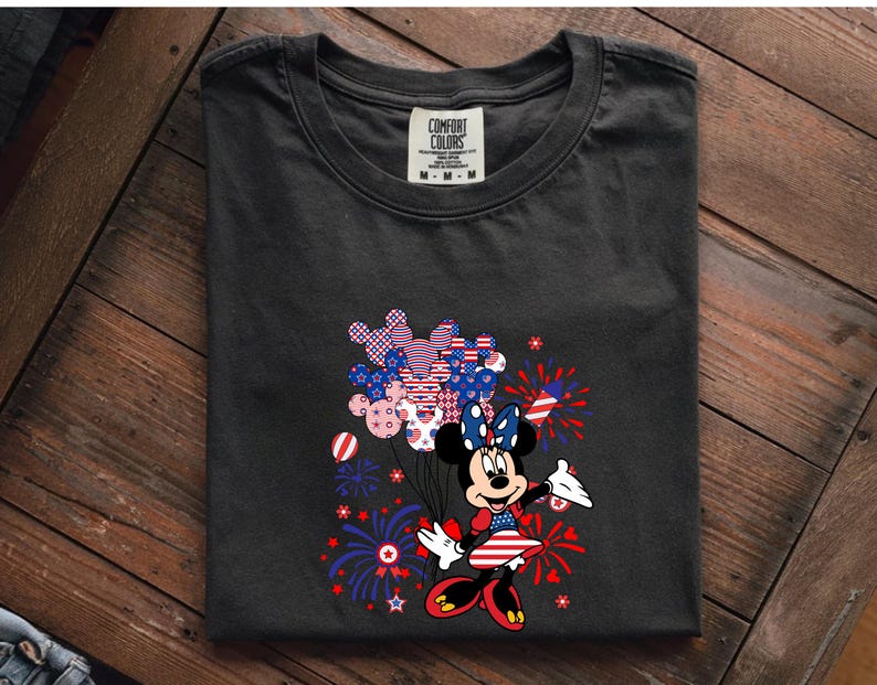 K&ouml;nnte beinhalten: Ein anthrazitfarbenes T-Shirt mit einer Grafik von Minnie Maus in einem rot-wei&szlig;-blauen Outfit. Sie h&auml;lt ein B&uuml;ndel Mickey-f&ouml;rmiger Luftballons mit patriotischen Mustern, umgeben von Illustrationen roter und blauer Feuerwerke.