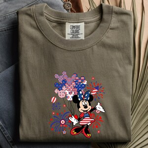 K&ouml;nnte beinhalten: Ein khakifarbenes T-Shirt mit einer Grafik einer Zeichentrickmaus in einem patriotischen Kleid, die ein B&uuml;ndel Luftballons in Form von Mausohren mit rot-wei&szlig;-blauen Mustern h&auml;lt, umgeben von Illustrationen von Feuerwerksk&ouml;rpern.